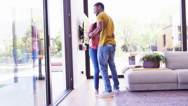 Photo shows an African American couple joyfully dancing near a large window at their home, expressing a moment of happiness and connection. Their body language showcases intimacy and comfort in a domestic setting. Ideal for use in content related to romance, modern lifestyles, or home living spaces.