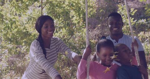 Parents pushing children on rope swing under sunlit canopy, family bonding outdoors