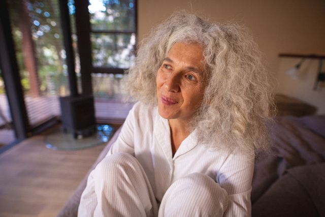 Senior woman with white hair sitting on a sofa in a living room, smiling and looking content. Natural light fills the room, creating a cozy and peaceful atmosphere. Ideal for use in articles or advertisements related to retirement, senior living, mental well-being, and home lifestyle.