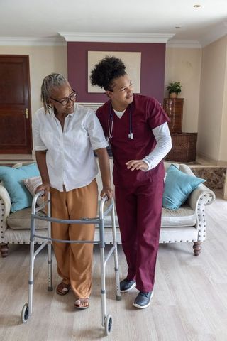 Young caregiver in burgundy scrubs assisting senior african american woman using walker at home