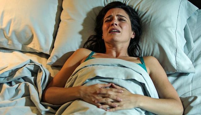 Woman lying in bed clutching abdomen and grimacing with pain at night