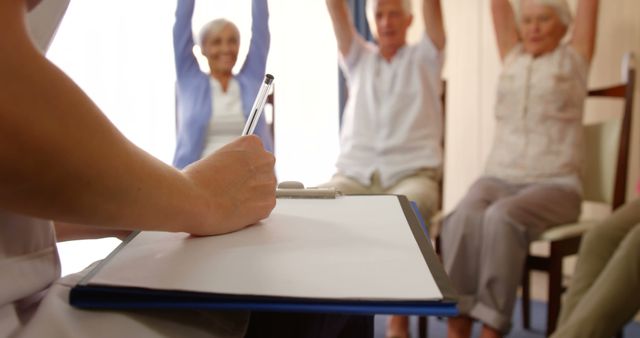 A group of seniors is participating in a group exercise session. They are seated with their arms raised, engaging actively in physical therapy. A healthcare professional is in the foreground, taking notes on a clipboard. This image can be used for promoting senior fitness programs, rehab centers, healthcare services for the elderly, and wellness initiatives.