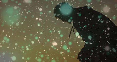 Hooded man leaning and bowing head amid falling bokeh lights and snow, moody silhouette