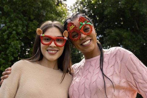 Diverse friends wearing festive novelty glasses outdoors smiling in knit sweaters