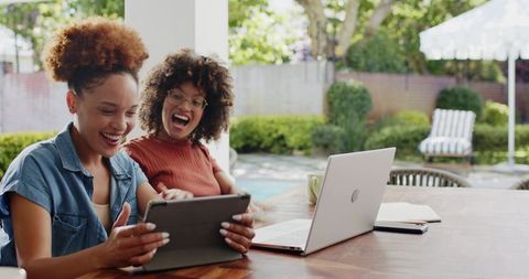 Laughing African American friends using tablet and laptop on sunny poolside patio