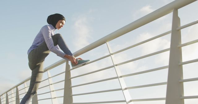 A young Muslim woman in a hijab is stretching her leg on a railing outdoors under a clear sky. She is wearing modern sportswear and appears to be preparing for a run or exercise routine. This image is ideal for illustrating themes of women's empowerment, fitness, health and wellness, diverse active lifestyles, and promoting inclusivity in sports. It can be used in blogs, fitness articles, health and wellness websites, or promotional material for athletic apparel.