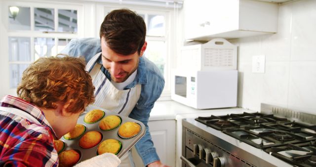 Father and child enjoying quality time baking muffins in a bright, modern kitchen. Great for portraying family bonding, parent-child activities, home life, and culinary interests. Perfect for advertisements, articles on parenting or cooking, and promotional materials for kitchen appliances or baking products.