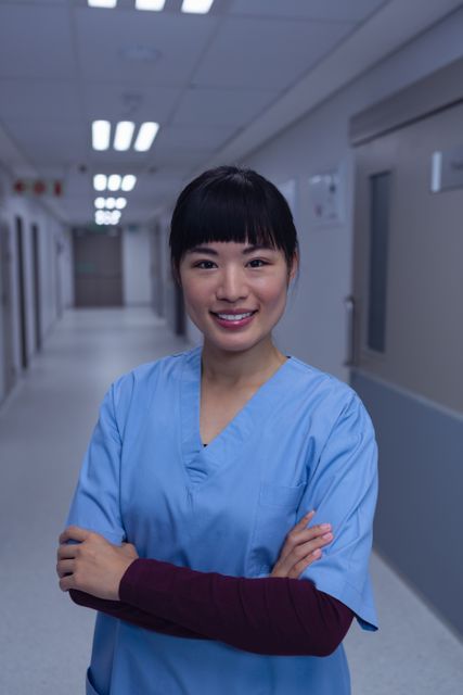 Confident female surgeon standing in a hospital corridor, smiling and with arms crossed. Ideal for use in healthcare-related articles, hospital websites, medical brochures, or health profession promotional materials.