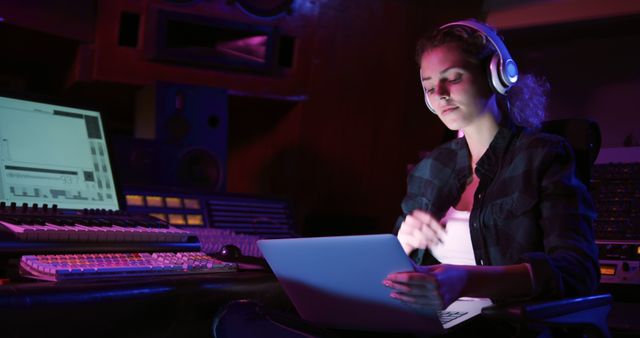 Young female music producer working on a laptop in a dimly lit studio at night, wearing headphones. Engaged in composing and mixing music, surrounded by professional audio equipment and screens. Ideal for concepts related to modern music production, digital content creation, professional sound engineering, and creative workflows in technology.
