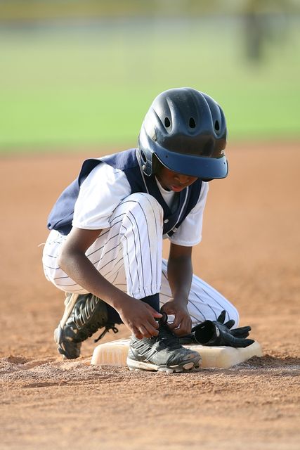 Image showing young baseball player kneeling at second base while tying shoelaces during game. Useful for illustrating youth sports, determination, preparation, training, and focus in athletics.