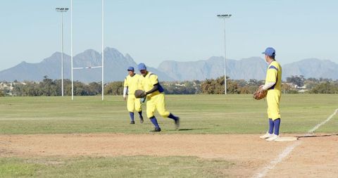 Baseball teammates fielding on grass diamond in yellow uniforms with mountain backdrop
