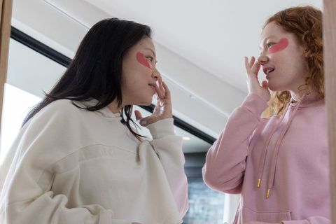Two friends applying heart-shaped eye patches in bright bathroom sharing skincare ritual