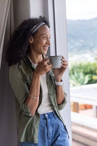 African american woman holding mug and gazing through sliding door, morning calm