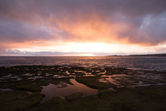 Image showcases a dramatic sunset over a rocky seashore with vivid skies and reflections on the water pools. Ideal for use in travel brochures, websites, and nature-themed inspirational content. Can be perfect for wallpapers or background images that project tranquility and beauty of natural landscapes.