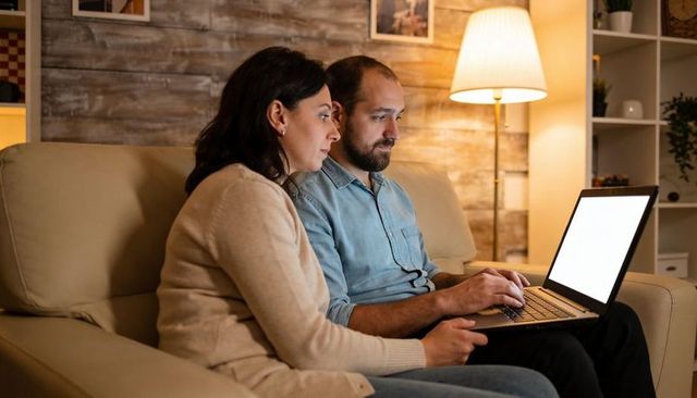 Couple browsing laptop on sofa at night in cozy living room, planning and streaming