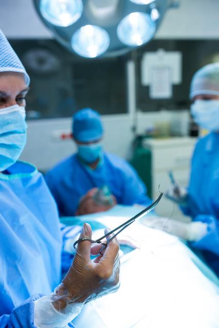 Surgeon examining surgical scissors in a well-lit operating room, with medical team members in the background. Ideal for use in healthcare, medical training, surgical procedures, hospital environments, and medical equipment promotions.
