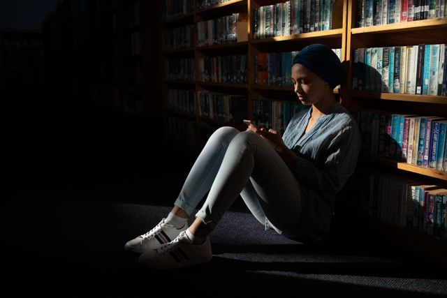 Side view of a biracial female student wearing a dark blue hijab studying in a library, sitting and holding a book in hands, reading it.