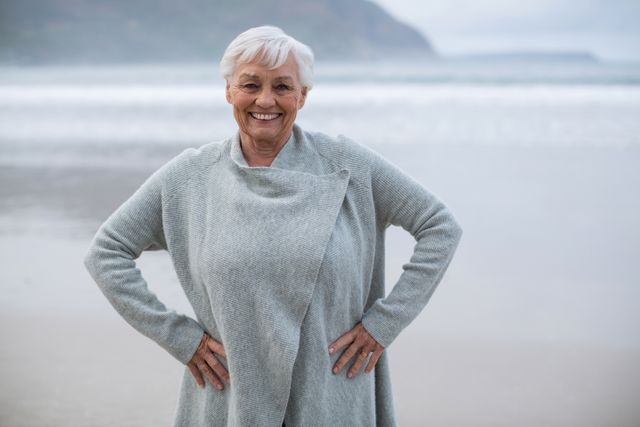 Elderly woman standing on beach with hands on hips, smiling warmly. Ideal for themes related to senior lifestyle, leisure, retirement, health, and well-being. Suitable for travel promotions, wellness campaigns, and social content celebrating active aging.