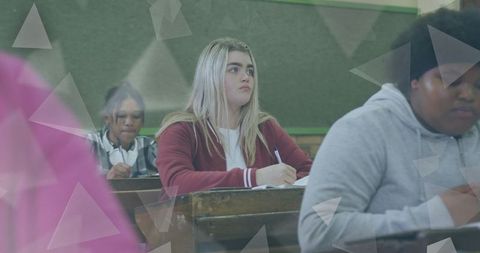 Blonde teen writing notes in classroom with diverse classmates studying at desks during lesson