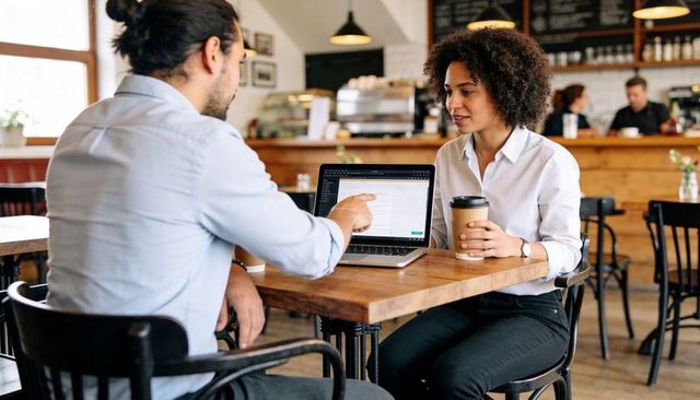 Colleagues discussing project over laptop while holding coffee in cozy urban cafe