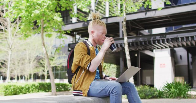 Young man sitting outside in an urban park, using laptop and enjoying coffee. Perfect for illustrating concepts of student life, outdoor work, relaxation, summer activities, and city parks.