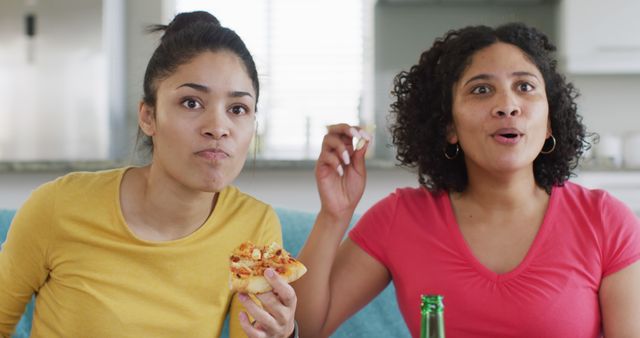 Two diverse happy female friends having beer and pizza watching tv at home, in slow motion. Free time, friendship and lifestyle concept.