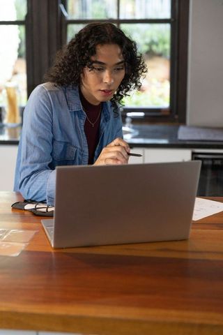 Nonbinary person working on laptop at wooden countertop holding stylus, home workstation lifestyle