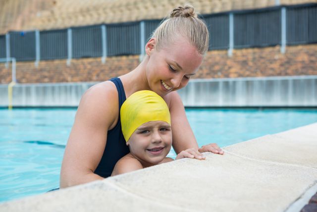 Female instructor and young boy relaxing at poolside in leisure center. The boy is wearing a yellow swim cap and smiling while the instructor supports him. Ideal for use in advertisements for swimming lessons, water safety programs, leisure centers, and summer activities.