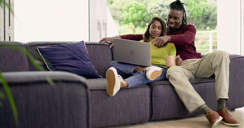 Diverse couple relaxing on sofa, sharing laptop and coffee in bright home living room