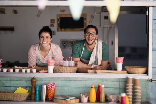 Two young adults, a waiter and a waitress, are smiling and leaning on the counter of a food truck. They are wearing aprons and appear friendly and approachable. The food truck is decorated with colorful items and has various condiments and utensils on the counter. This image can be used for promoting small businesses, street food events, customer service training, or entrepreneurship.