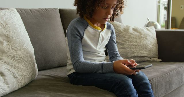 A young boy with curly hair, dressed in casual clothes, is sitting on a sofa inside a home, focused on using a smartphone. This image can be useful for illustrating concepts related to childhood and technology, digital interaction, home lifestyle, or family life.