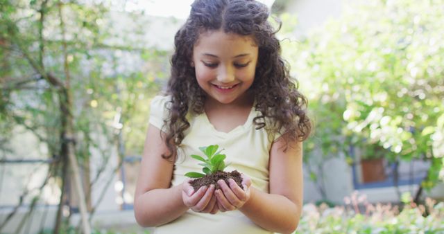 Smiling girl holding a young plant in her hands while standing in a garden. This image can be used for educational materials on environmental responsibility, promoting gardening activities for children, or illustrating sustainable living practices. Suitable for websites, articles, and campaigns focused on nature, outdoor activities, and childhood development.