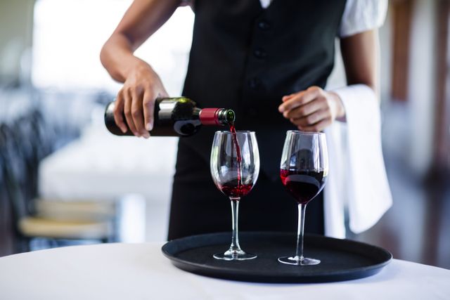Waitress pouring red wine into glasses on a serving tray in a restaurant. Ideal for illustrating concepts of hospitality, fine dining, professional service, and restaurant settings. Useful for marketing materials for restaurants, hospitality training, and dining experience promotions.