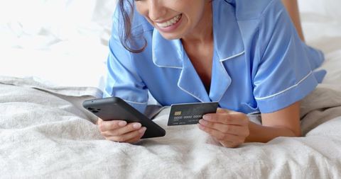 Happy woman shopping online with smartphone and credit card in bed