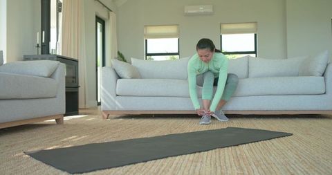 Mid-adult asian woman tying sneakers before home workout on bright living room mat
