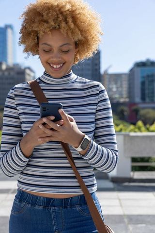 Smiling african american woman texting on smartphone on sunlit city rooftop terrace