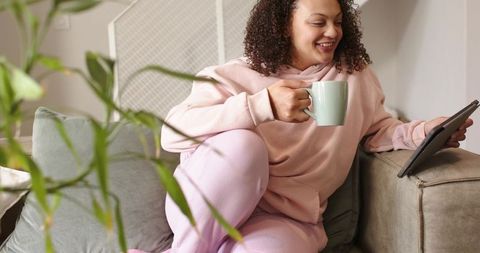 African american woman relaxing on couch in pale pink hoodie holding mint mug and tablet