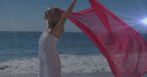 Windblown woman raising flowing red scarf over ocean shoreline in white strapless dress