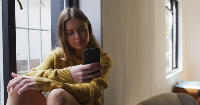 Young woman sitting by window using smartphone in cozy indoor setting - Download Free Stock Images Pikwizard.com