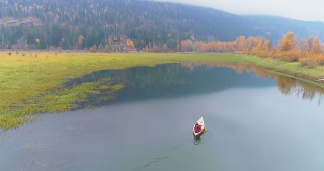 A lone individual paddling a canoe on a calm lake surrounded by autumn trees and forested hills. Ideal for promoting outdoor activities, travel, relaxation, and nature retreats. Can be used for articles or advertisements related to wilderness adventures, mental well-being, or seasonal transitions.