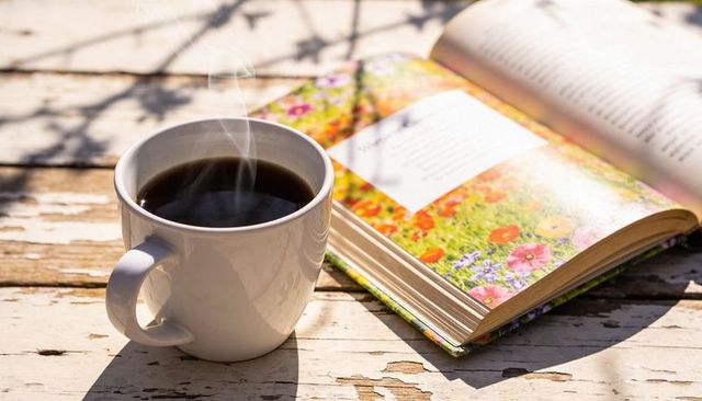 Steaming coffee mug on weathered wood table with open floral hardcover book in warm sunlight
