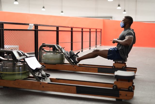 African american man wearing a facemask while working out on the rolling seat exercise machine. he is pulling the string close to his chest.