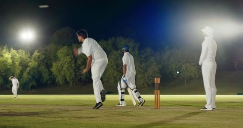 Diverse male cricketers playing night match under floodlights bowling and batting action