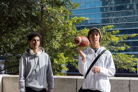Asian and african american teens spinning football on urban terrace with headphones and hoodies