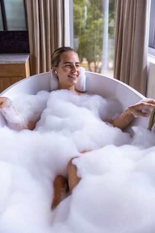 Woman relaxing in bubble bath by window with earbuds and towel headrest