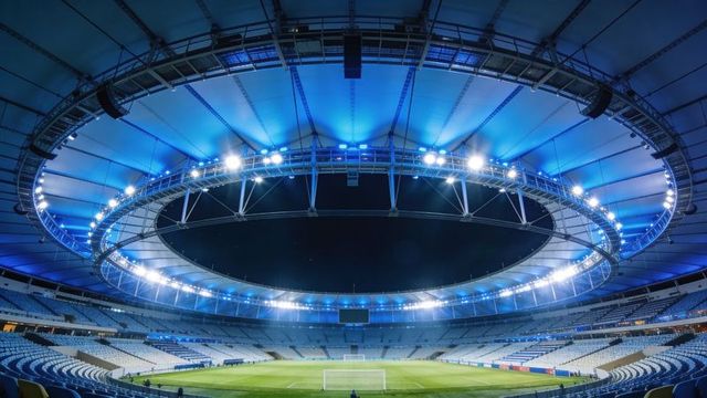 Roofed soccer stadium glowing with circular ring floodlights over central pitch at night