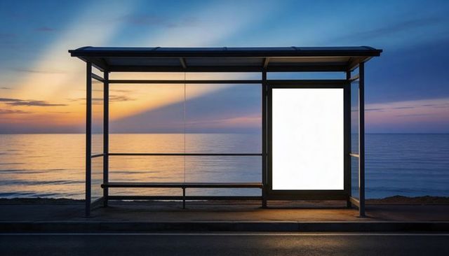 Glowing seaside bus shelter casting light from blank illuminated ad panel at sunset