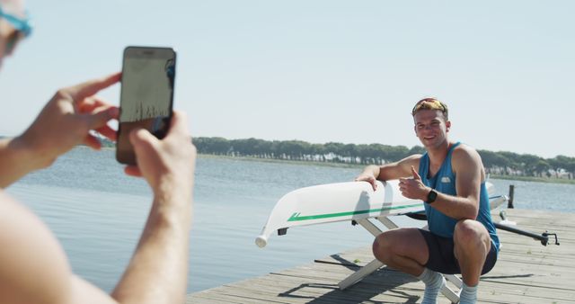 Caucasian male rower taking picture of happy teammate with thumb up by boat on jetty on sunny day. Communication, social media, summer, sport, rowing, hobbies and active lifestyle, unaltered.