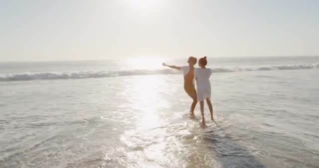 Young man and woman dancing and holding hands on a sunny beach with ocean waves crashing. Suitable for themes of romance, summer vacations, joy, and young love. Ideal for travel blogs, lifestyle content, advertisements, and vacation promotions.