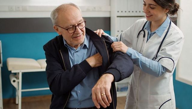Female physician helping senior man put on jacket during clinic visit, patient-centered care